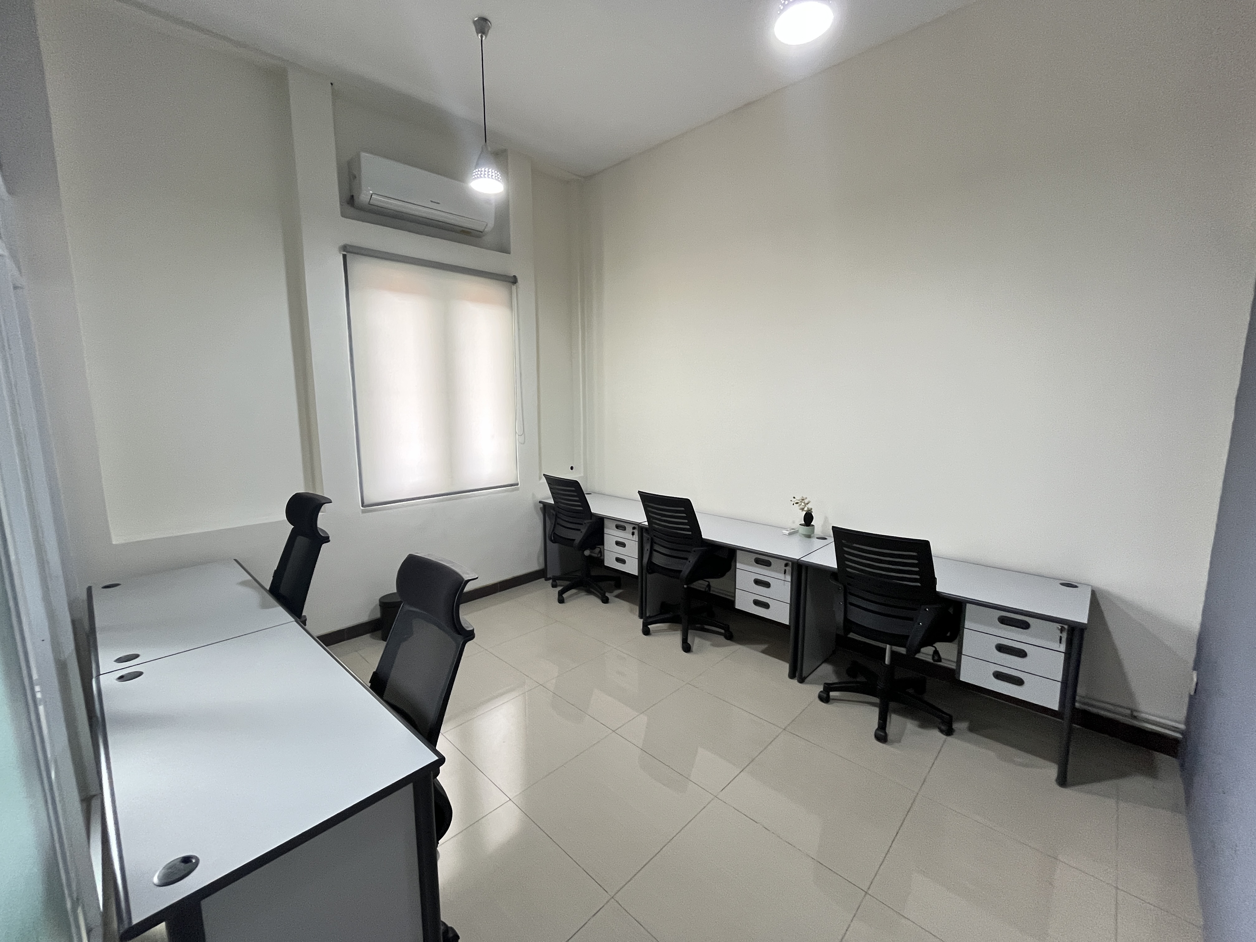 Private office with white desks with drawers, black chairs, a window with a roller blind, and an air conditioner